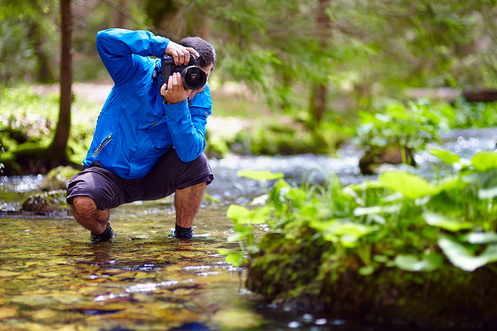 Professional,Nature,Photographer,Taking,Photos,By,The,River,Side Photographer crouched in woodland river in a blue jacket